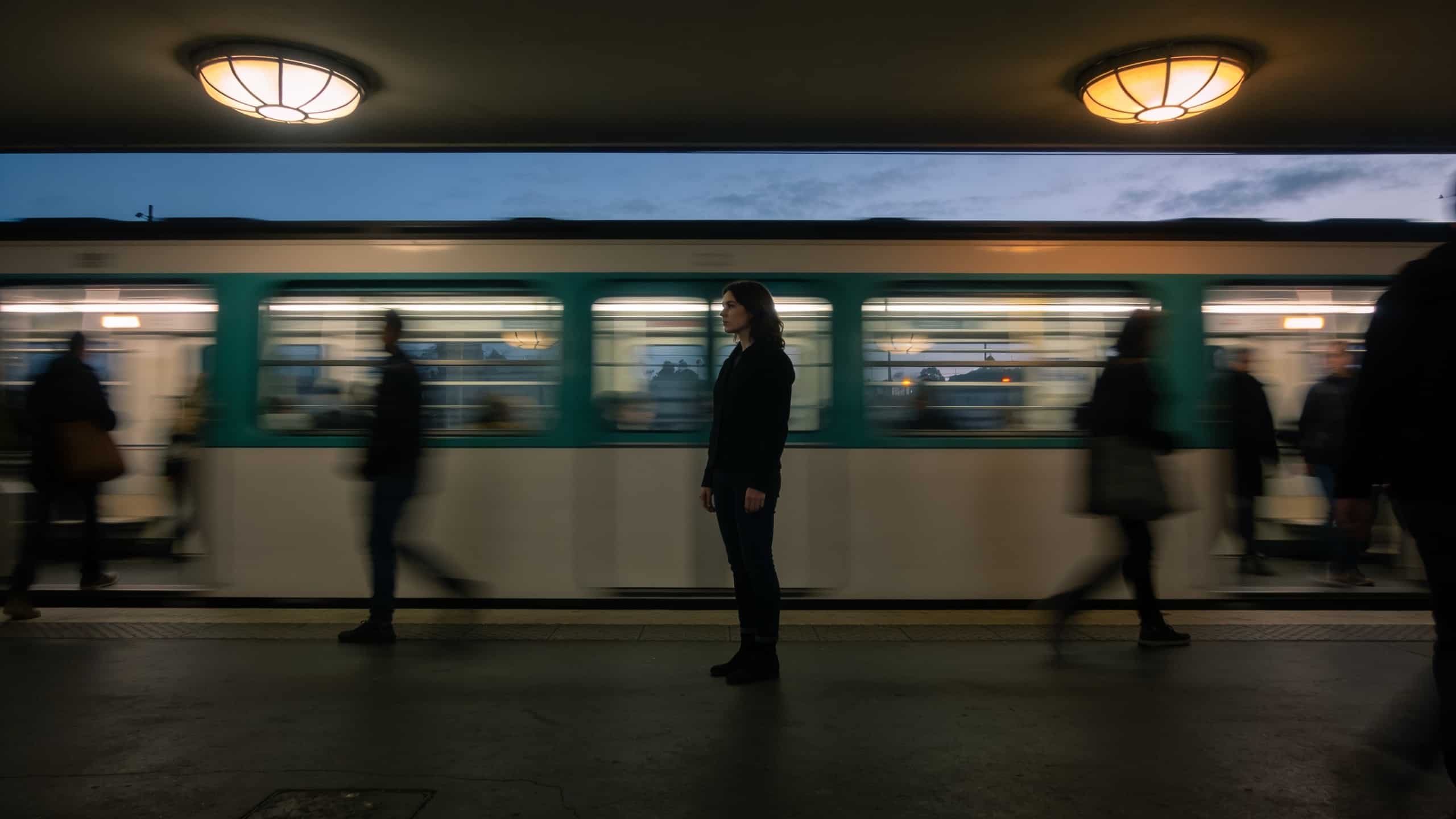 limerence — Outdoor/urban transitional space: an above-ground Metro platform at dusk, a single figure mid-frame standing s...