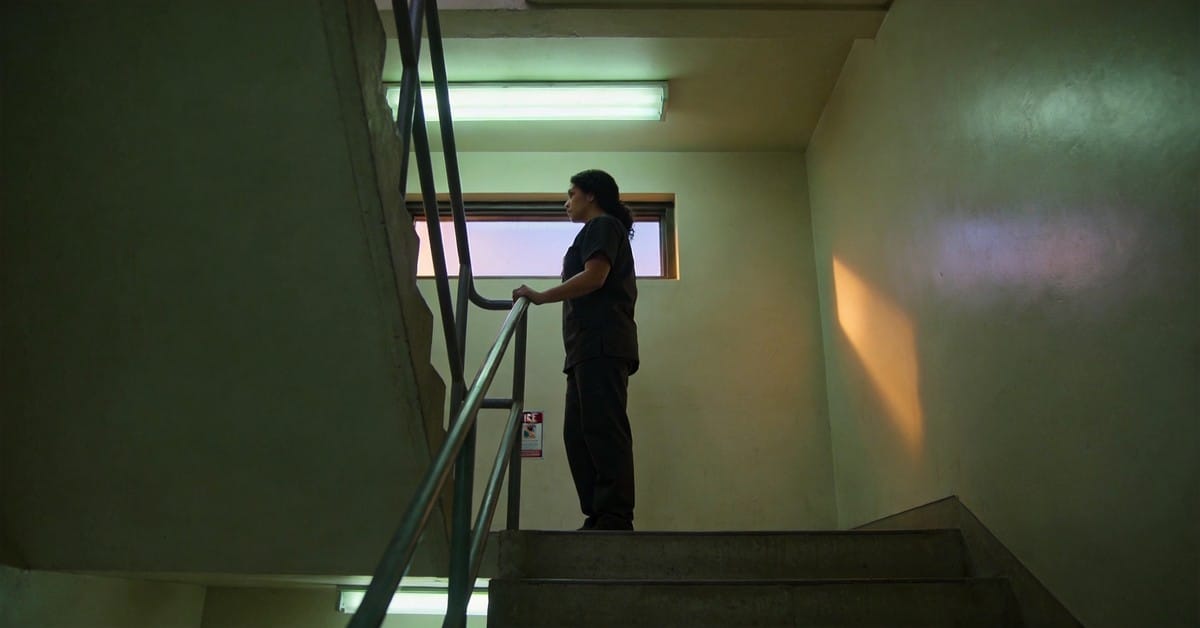 physician burnout — A figure in scrubs paused in a hospital stairwell landing between floors at dusk — institutional fluor...