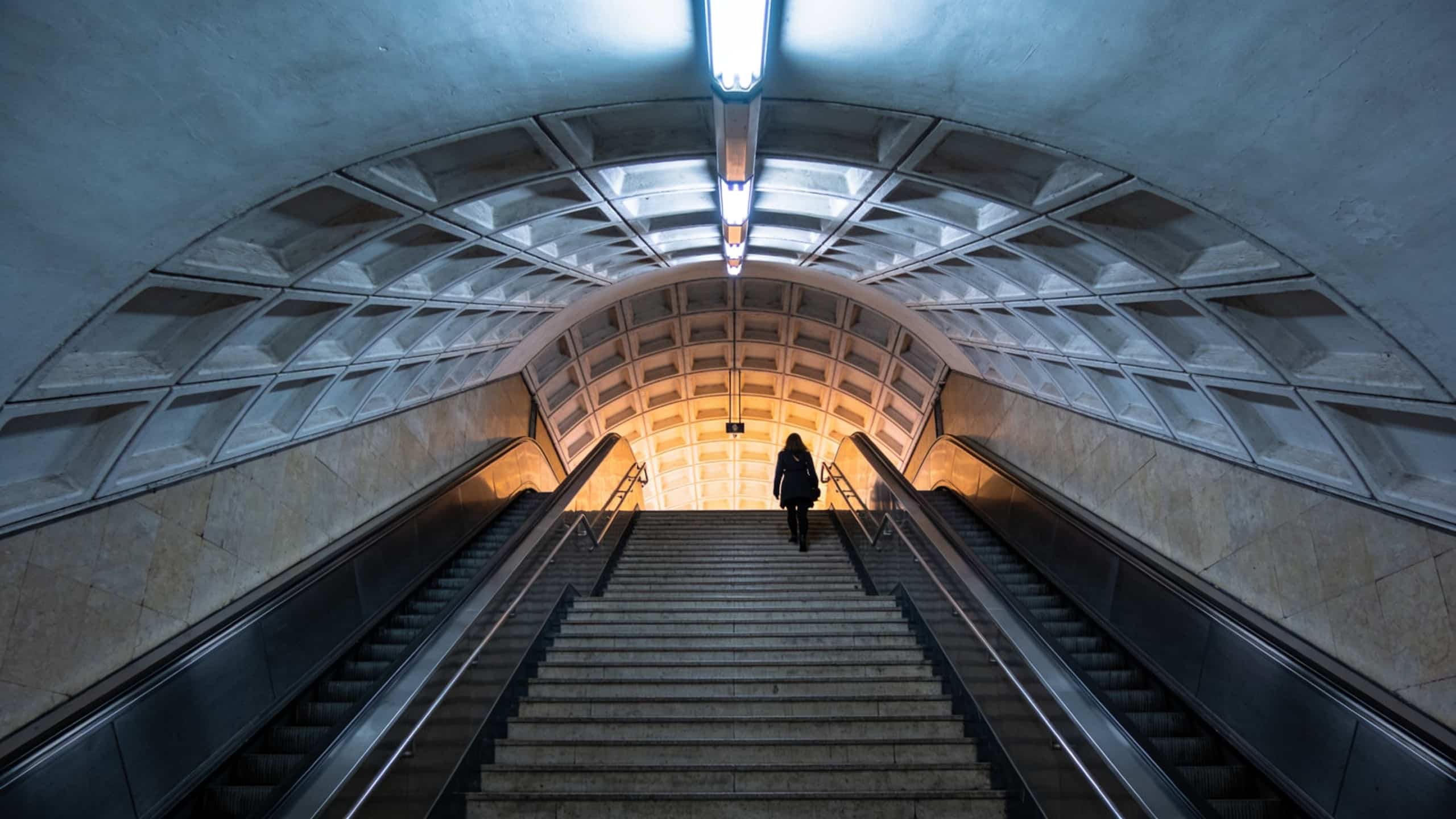A person climbs the stairs between two escalators under the coffered arch of a Metro station.